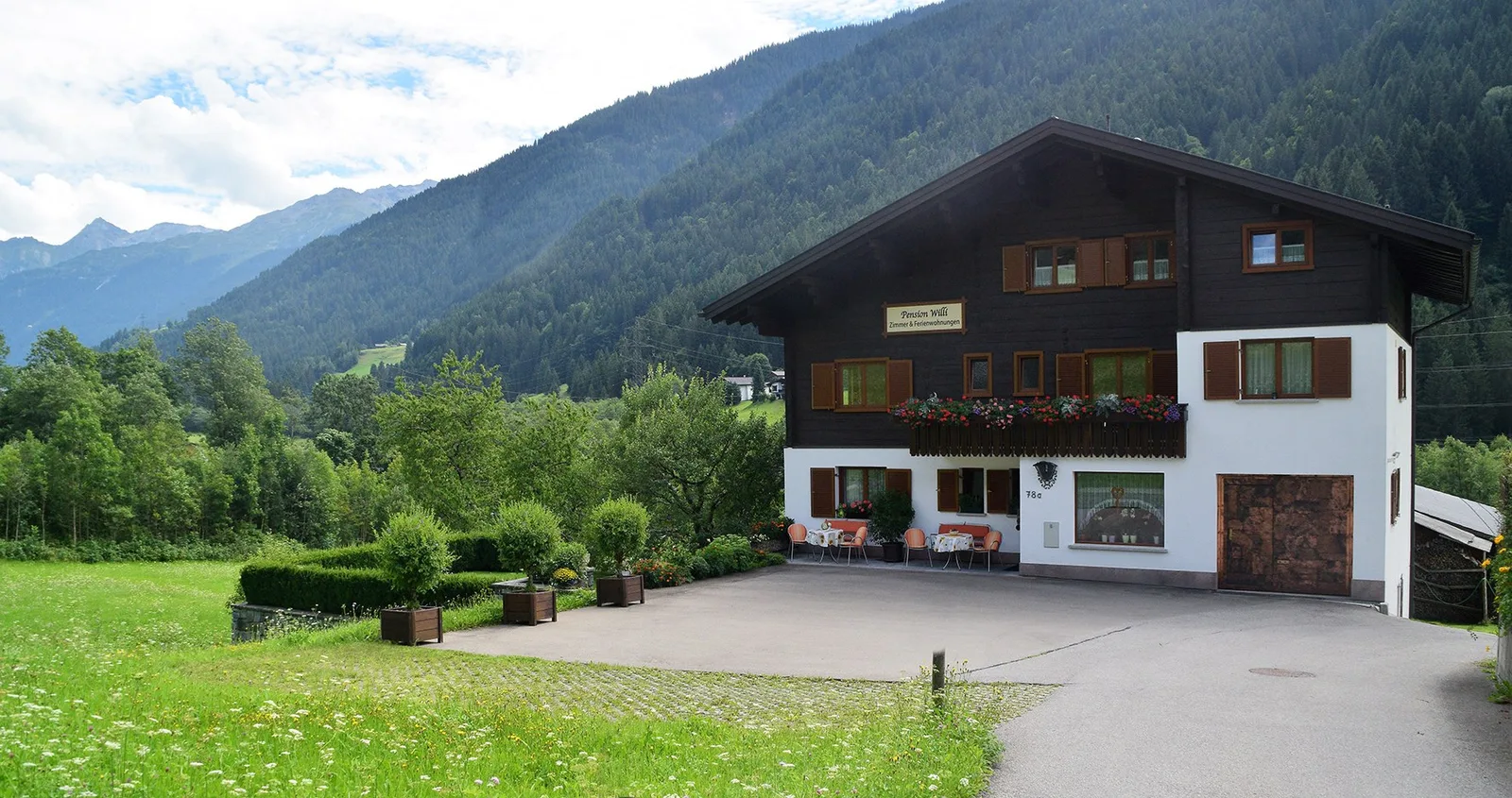 Das Haus der Pension Willi im Panorama, davor die Wiese, dahinter die Berge